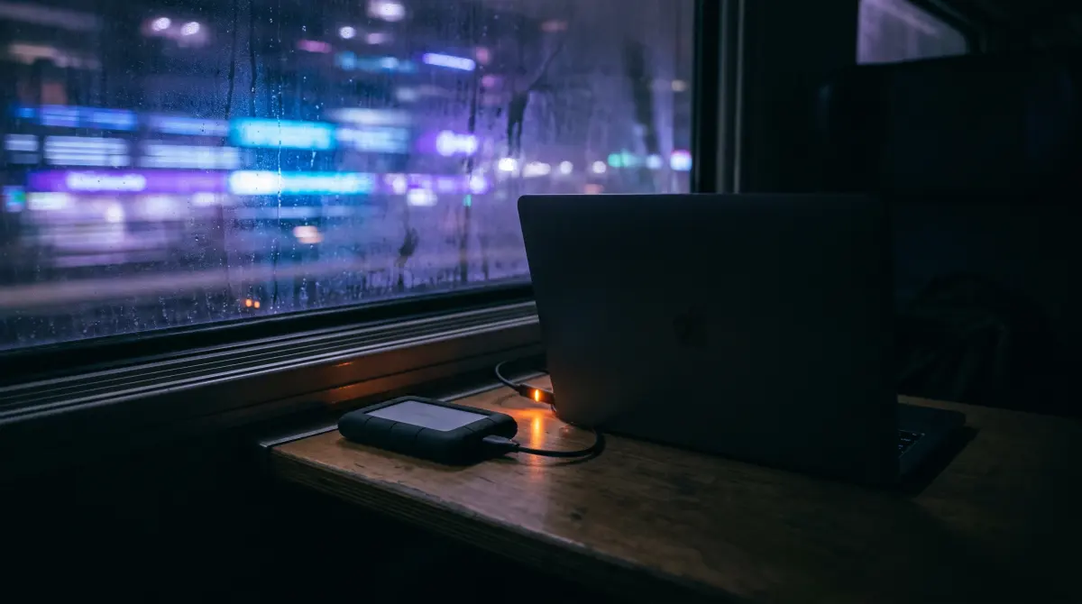 A laptop and portable drive on a night train table by a streaked window