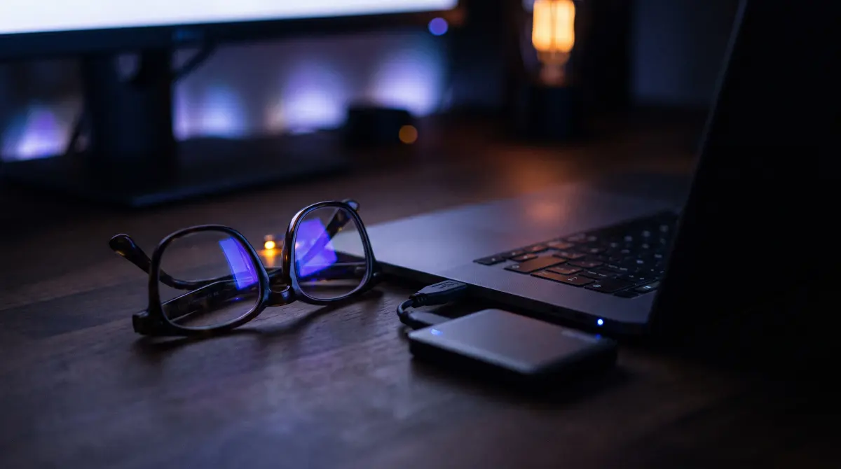 Glasses beside a laptop reflect the screen's blue glow, a portable drive plugged in nearby
